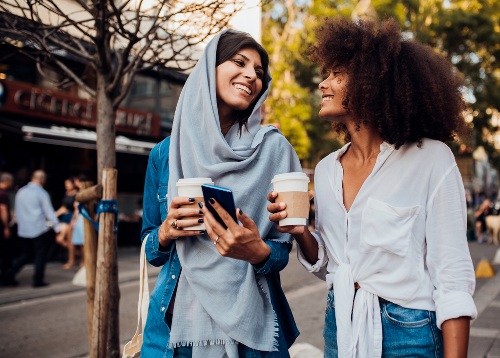 Two friends enjoying a hot girl walk. The woman on the left wears a slate blue headscarf while her friend shows off her long, natural black hair blowing in the breeze. Their smiles radiate the joy of sisterhood.