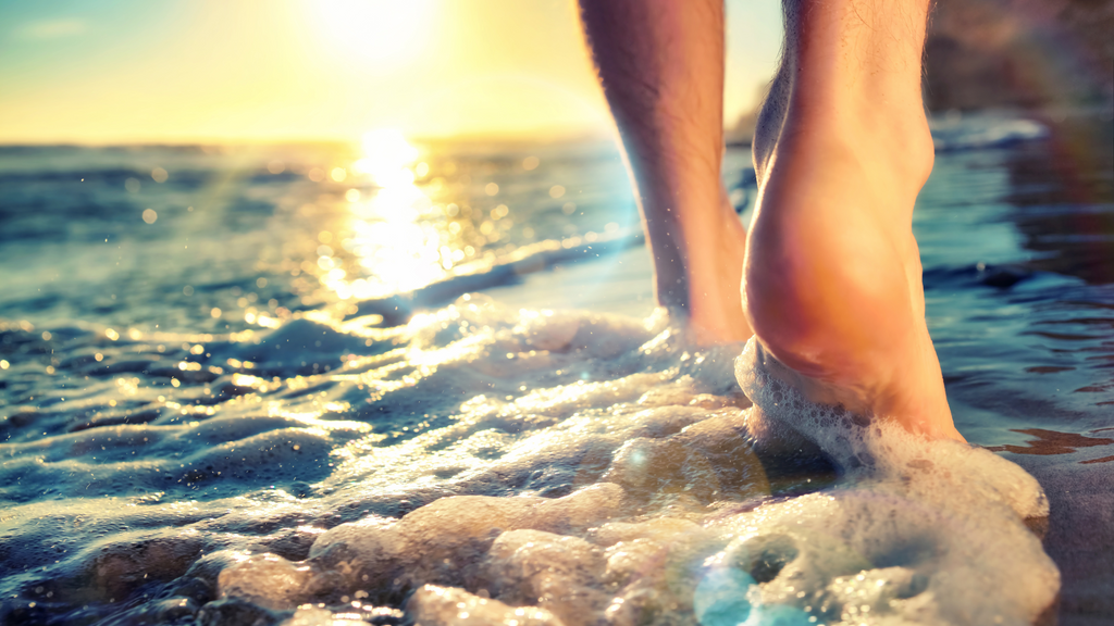 A pair of men's feet walk along a the beach with foamy waves carshing across their toes.